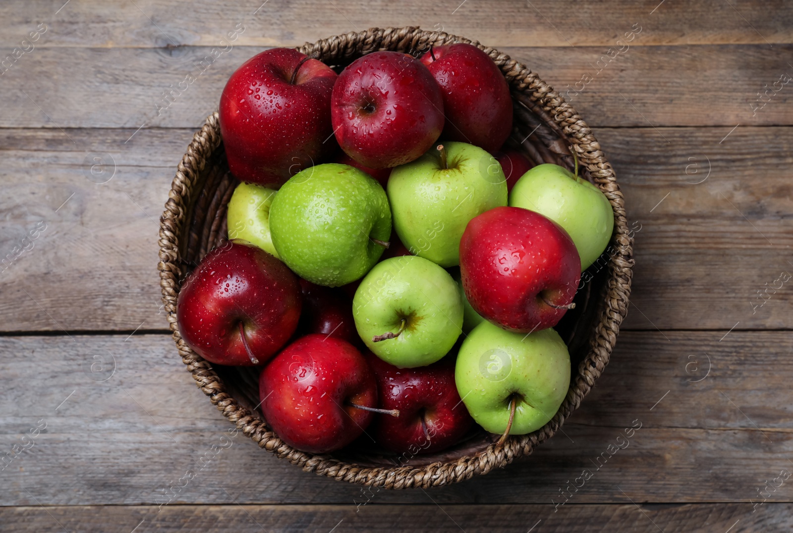 Photo of Fresh ripe green and red apples with water drops in wicker bowl on wooden table, top view