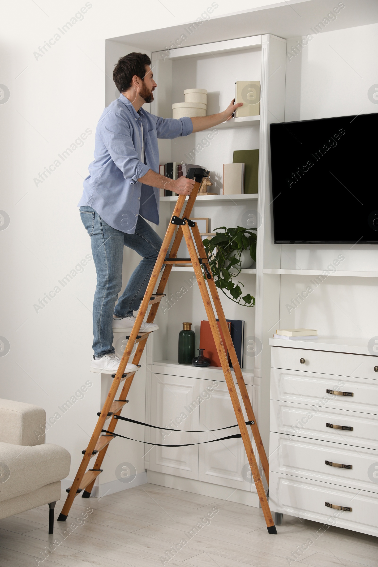 Photo of Man on wooden folding ladder taking book from shelf at home