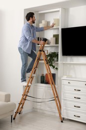 Photo of Man on wooden folding ladder taking book from shelf at home