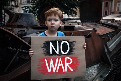 Photo of Sad boy holding poster No War near broken military tank outdoors