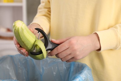 Photo of Woman peeling fresh zucchini above garbage bin indoors, closeup