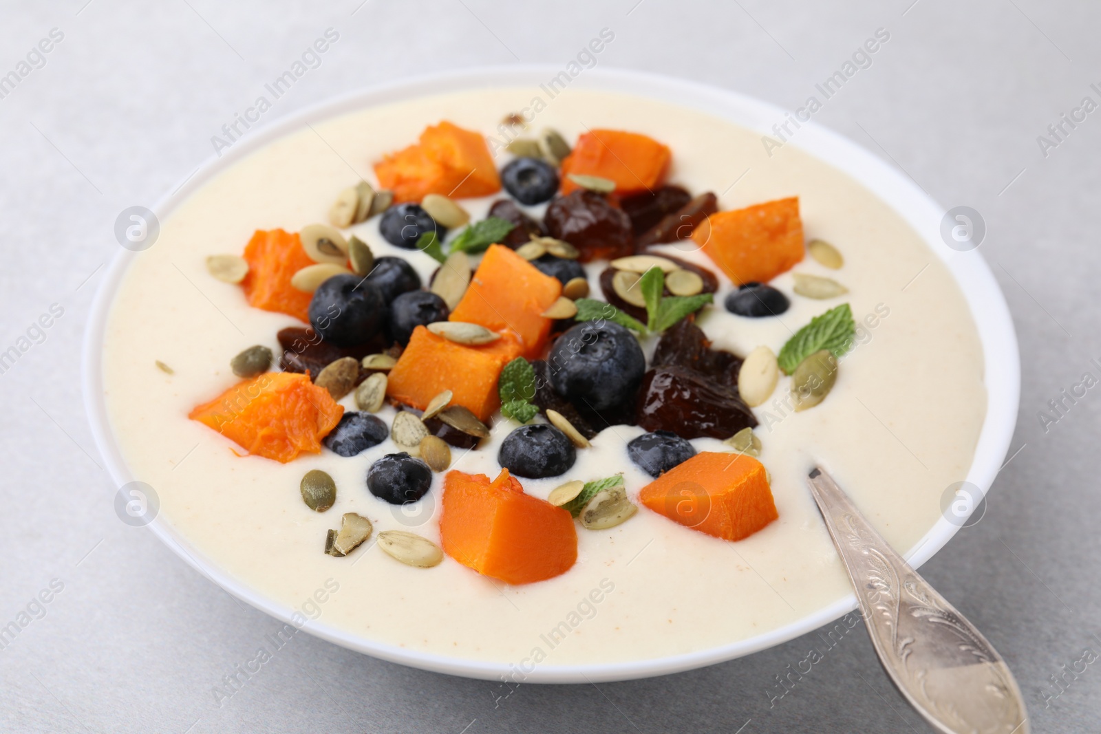 Photo of Delicious semolina pudding with blueberries, dates, pumpkin, mint and spoon in bowl on white table, closeup