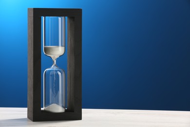 Photo of Hourglass with light green flowing sand on white table against blue background. Space for text