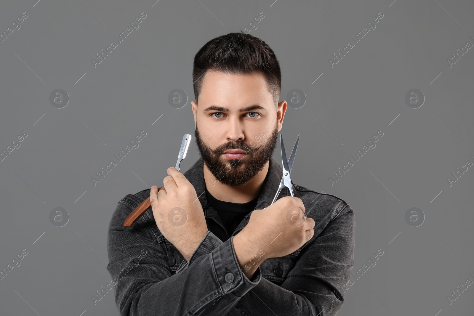 Photo of Handsome young man with mustache holding blade and scissors on grey background