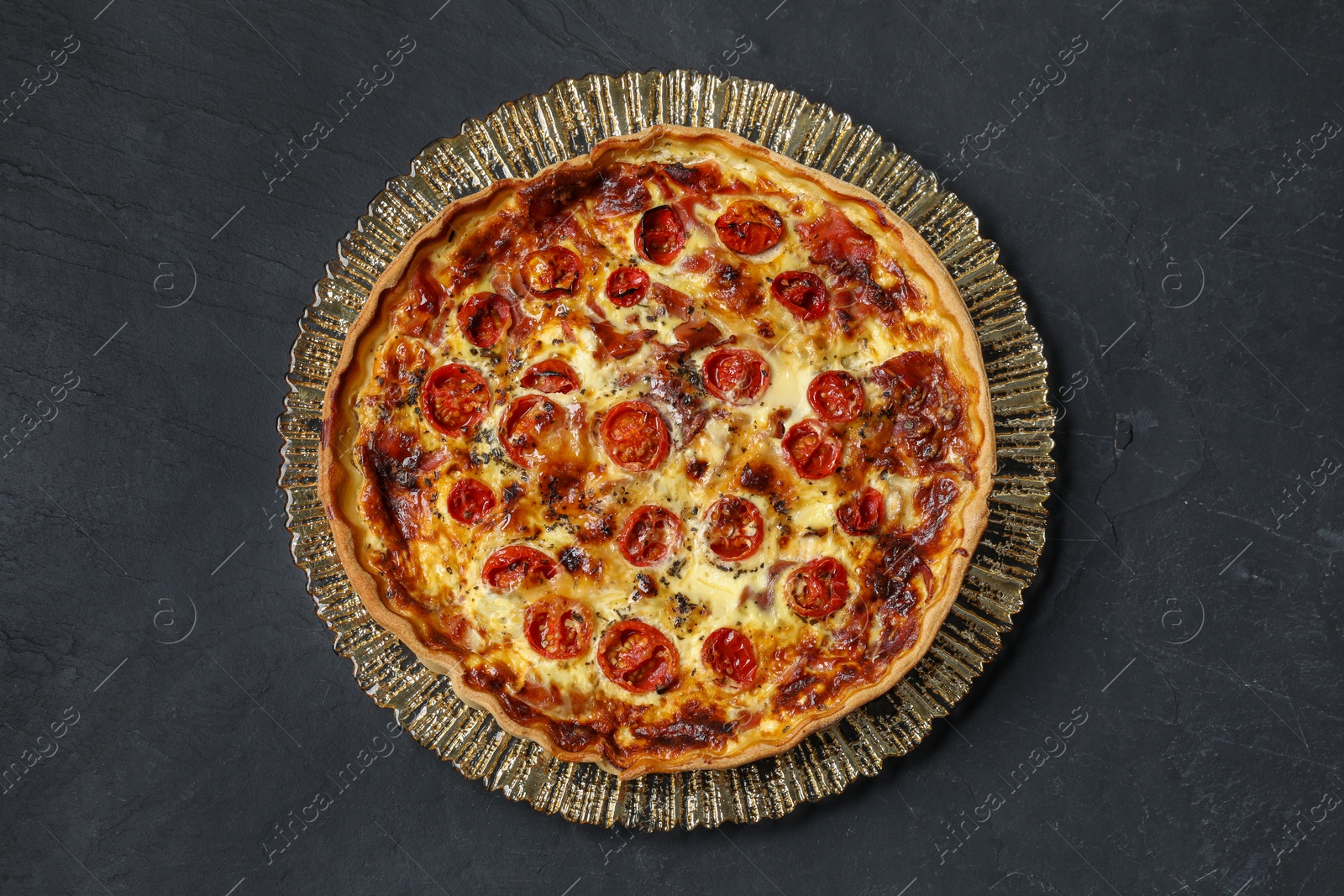 Photo of Delicious homemade quiche with prosciutto and tomatoes on black textured table, top view