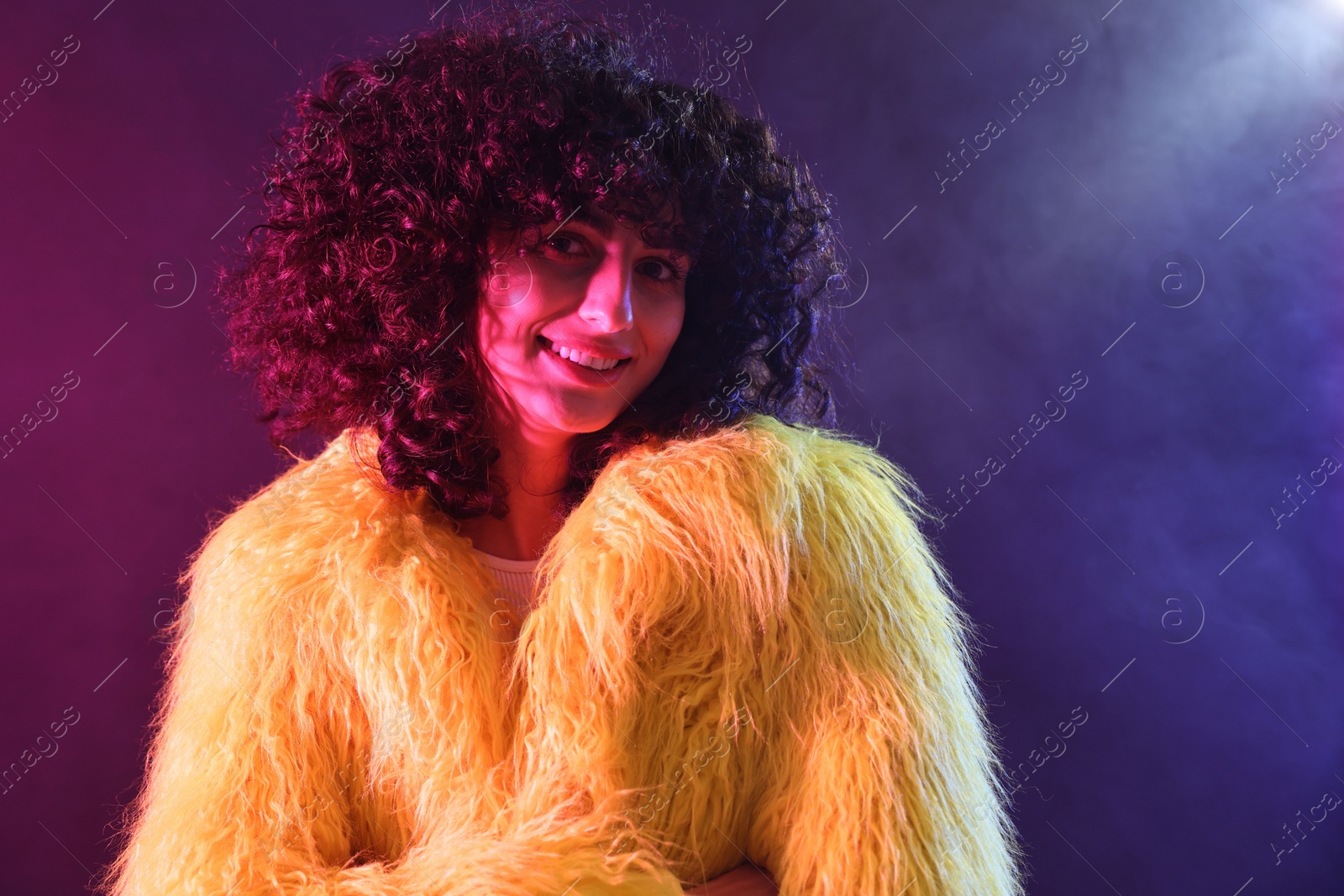 Photo of Beautiful young woman in yellow fur coat on color background in neon lights and smoke