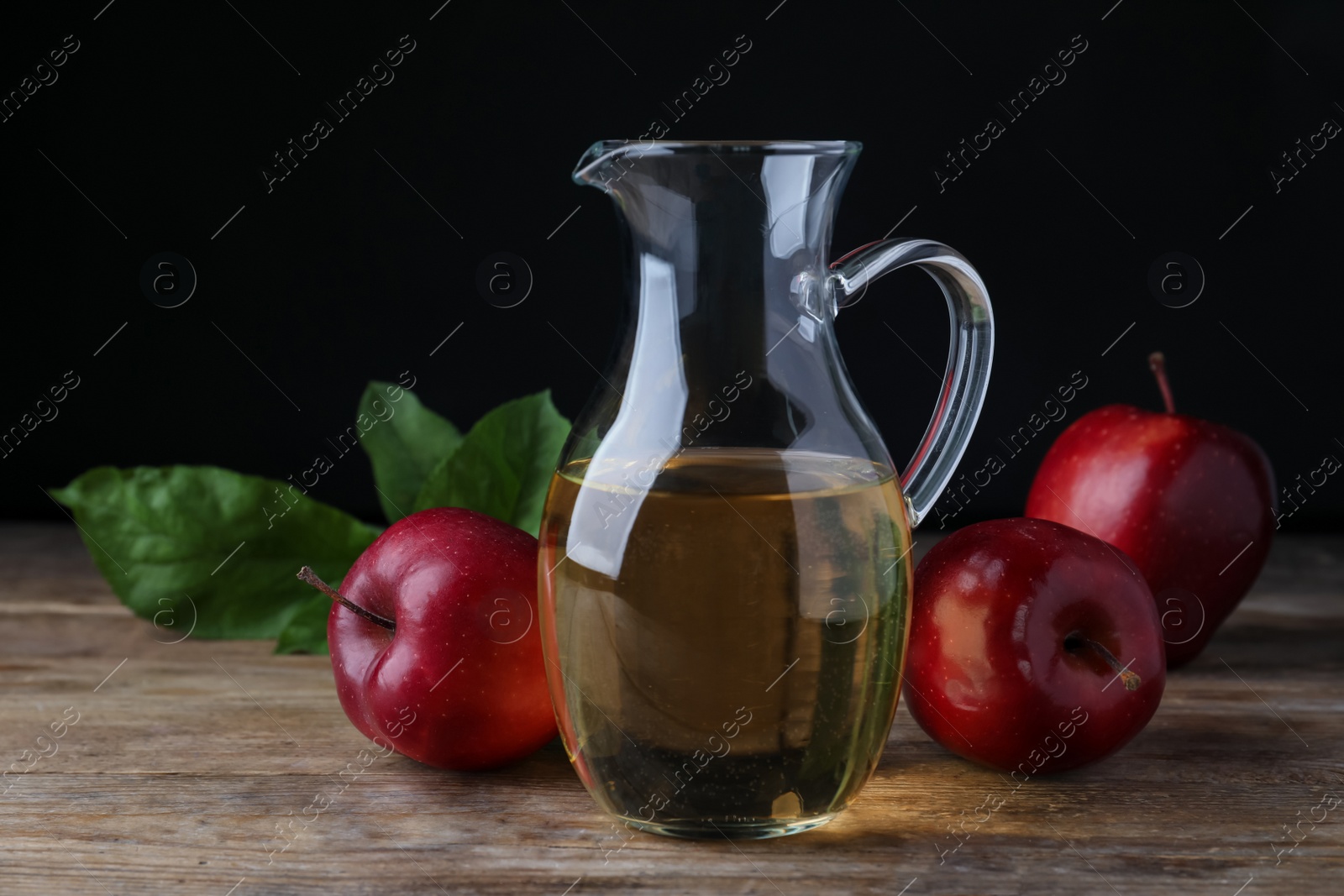 Photo of Jug of tasty juice and fresh red apples on wooden table against black background