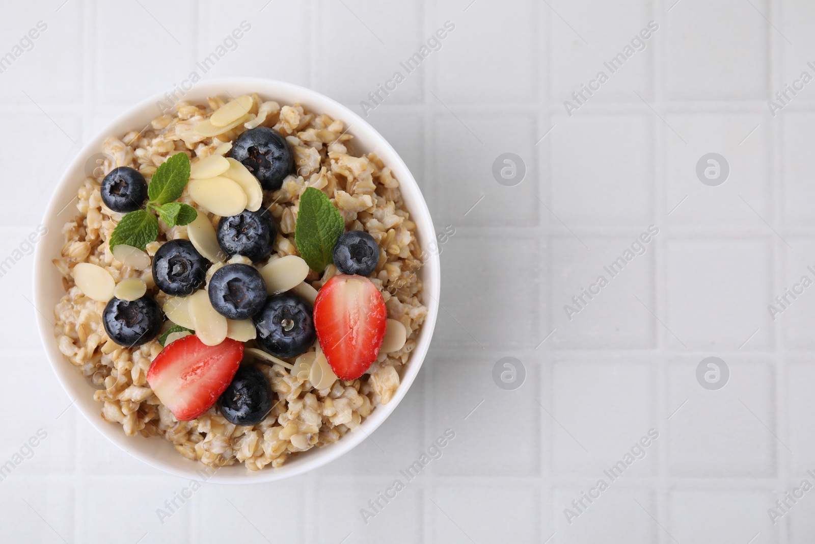 Photo of Tasty oatmeal with strawberries, blueberries and almond petals in bowl on white tiled table, top view. Space for text