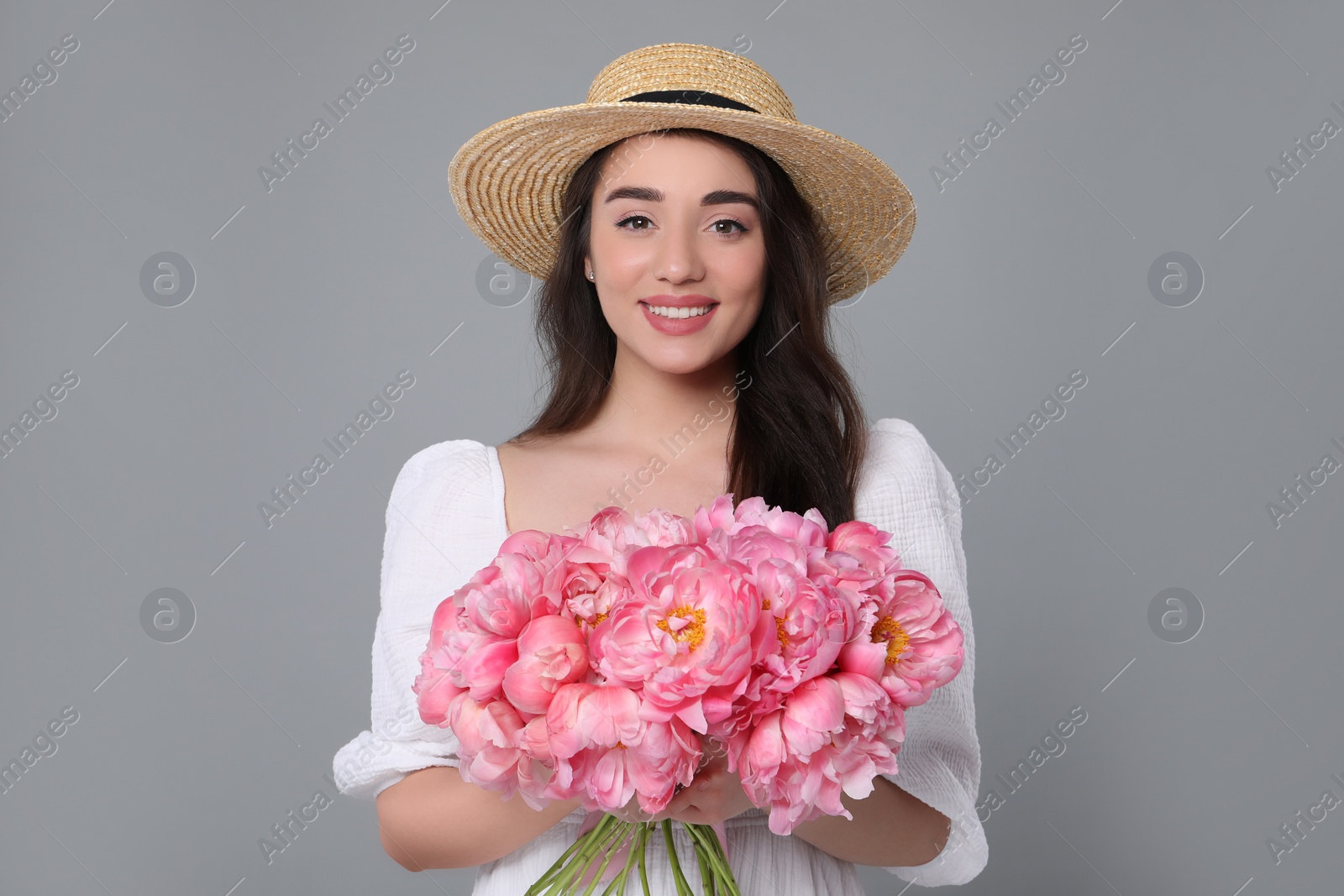 Photo of Beautiful young woman in straw hat with bouquet of pink peonies against grey background