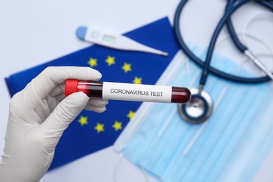 Photo of Doctor holding sample tube with label Coronavirus Test above medical items and European Union flag, closeup