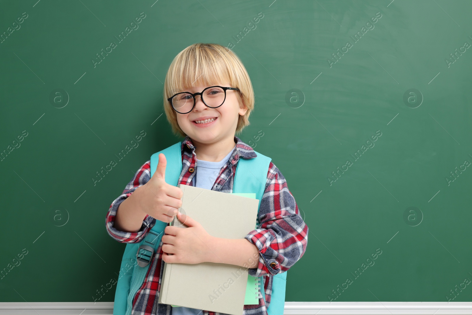 Photo of Happy little school child with notebooks showing thumbs up near chalkboard. Space for text