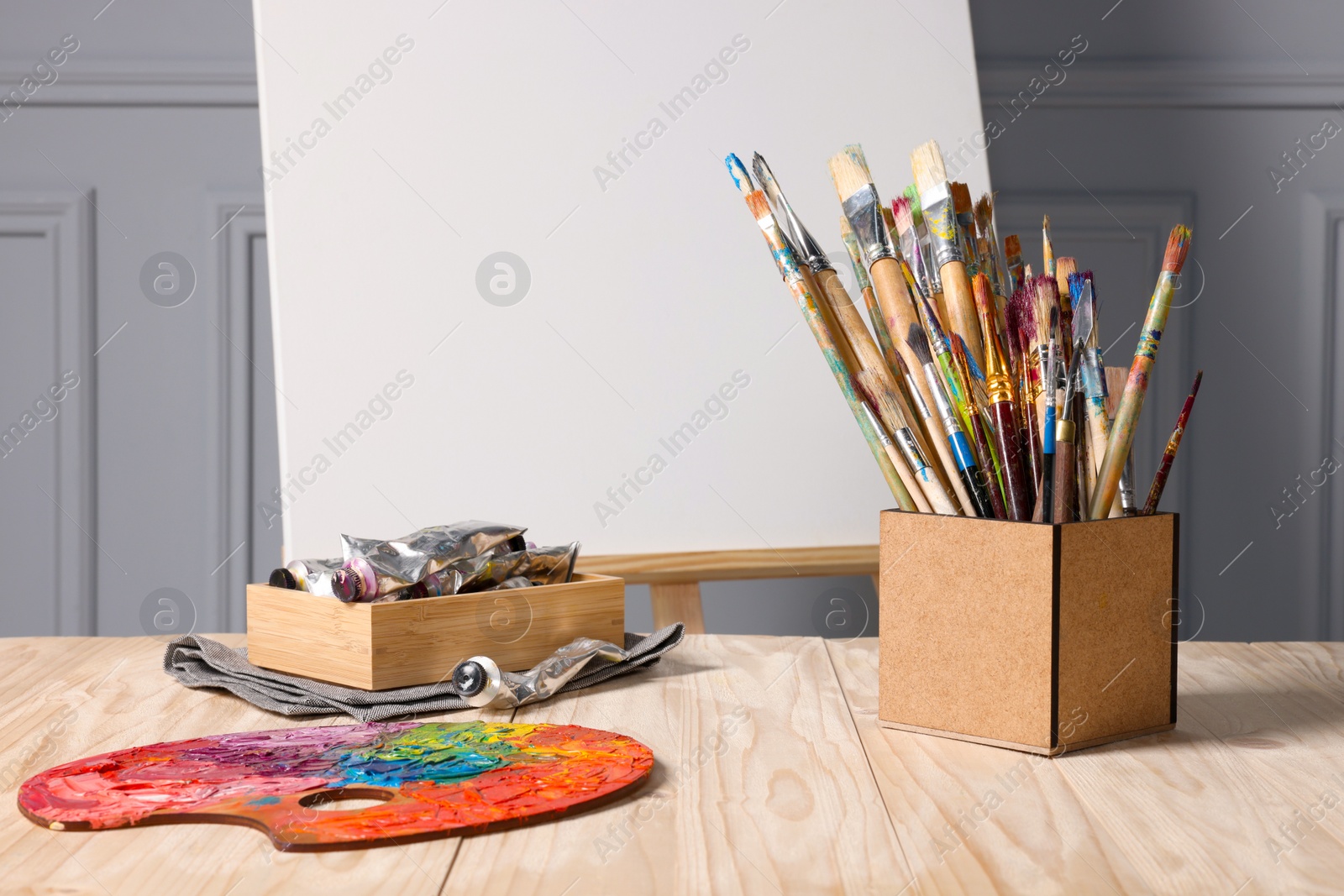 Photo of Easel with blank canvas and different art supplies on wooden table near grey wall