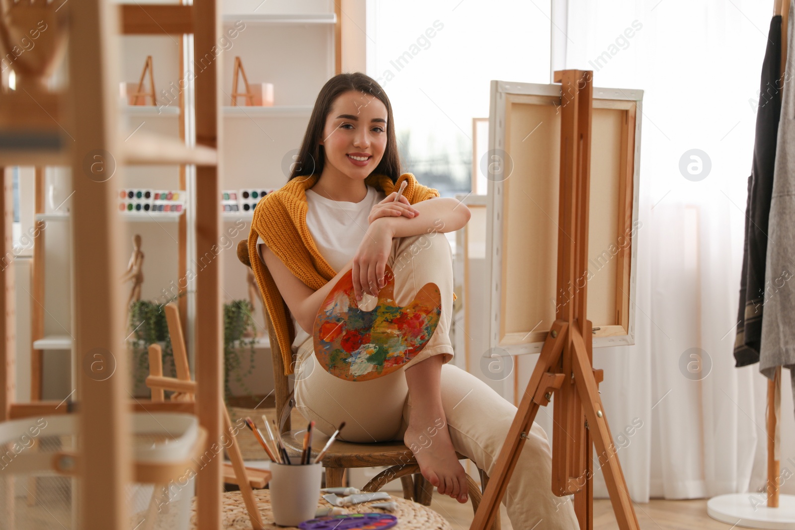 Photo of Beautiful young woman with drawing tools near easel at home