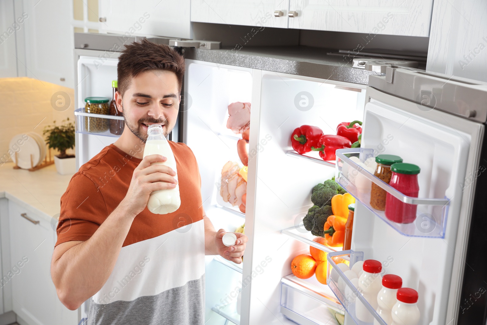 Photo of Young man drinking milk near open refrigerator in kitchen