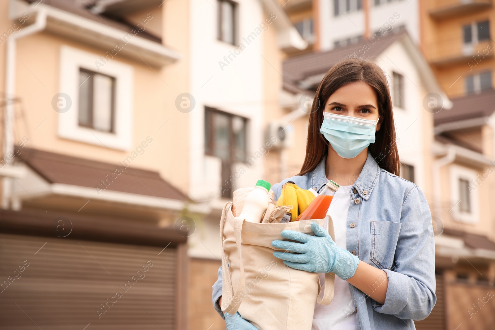 Photo of Female volunteer in protective mask and gloves with products on city street. Aid during coronavirus quarantine