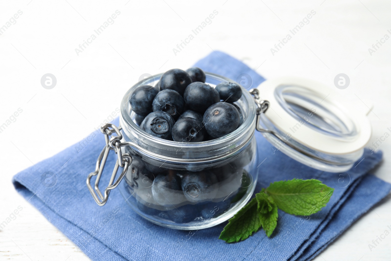 Photo of Glass jar of tasty fresh blueberries, mint leaves and fabric on white wooden table