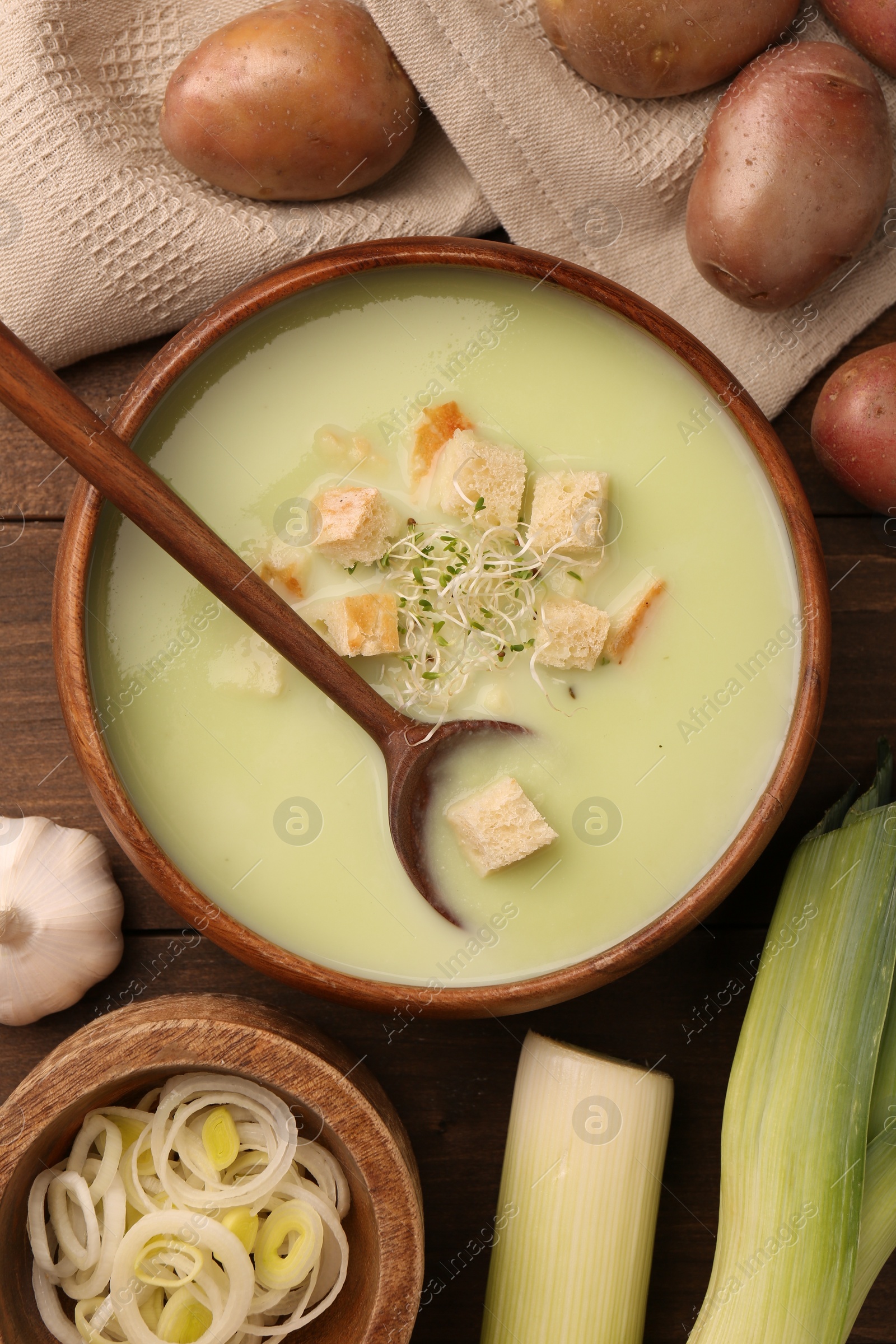 Photo of Bowl of tasty leek soup, spoon and ingredients on wooden table, flat lay