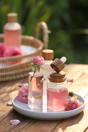 Photo of Bottles of rose essential oil and flowers on wooden table outdoors
