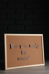 Photo of Cork board with phrase Are You Ready For Exams? on wooden table near black brick wall