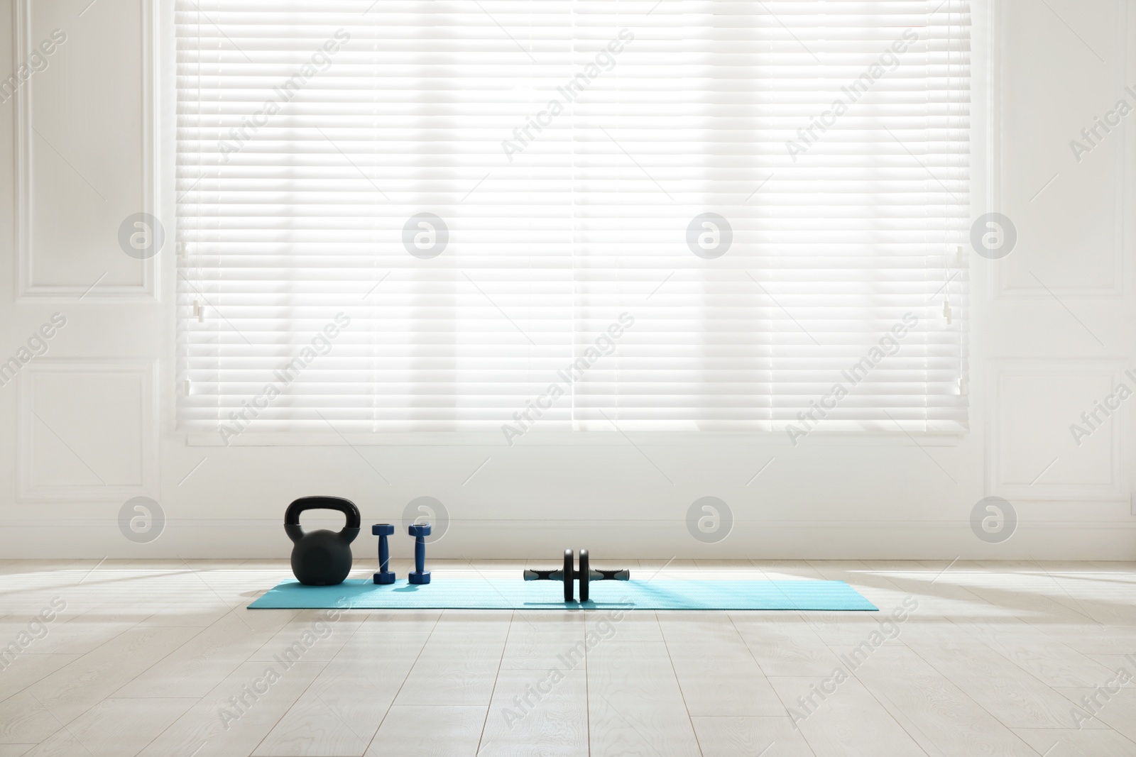 Photo of Exercise mat, ab roller, dumbbells and kettlebell near window in spacious room