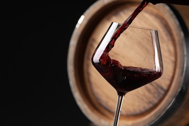 Pouring red wine into glass near wooden barrel against black background, closeup. Space for text