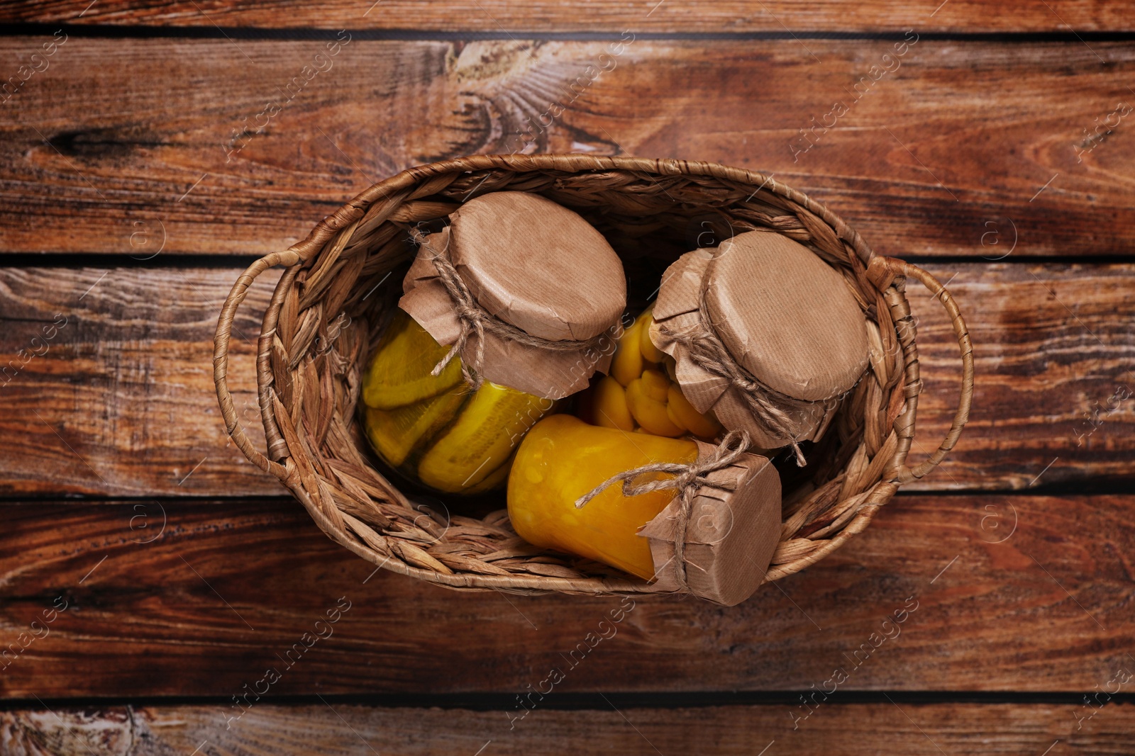 Photo of Wicker basket with many jars of different preserved products on wooden table, top view