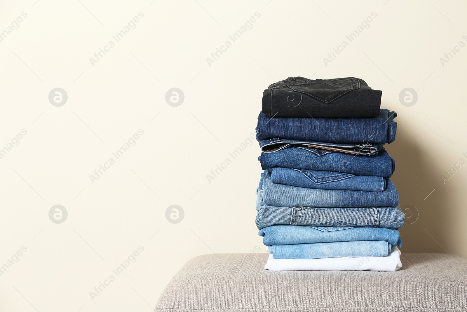 Photo of Stack of different jeans on ottoman against light background. Space for text