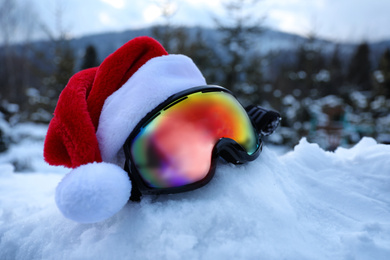 Photo of Stylish ski goggles with Santa hat on snow outdoors. Winter sport equipment