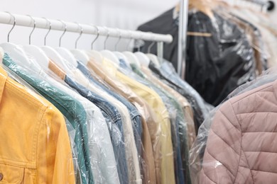 Photo of Dry-cleaning service. Many different clothes in plastic bags hanging on rack indoors, closeup