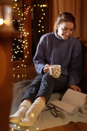 Photo of Woman with cup of hot beverage reading book at home in winter evening