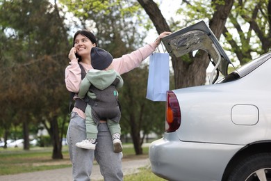 Photo of Mother holding her child in sling (baby carrier) while closing car trunk outdoors