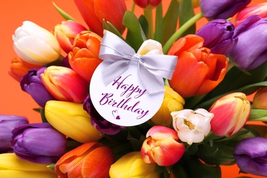 Image of Beautiful bouquet of tulip flowers with Happy Birthday card on orange background, closeup