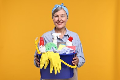 Photo of Happy housewife holding bucket with cleaning supplies on orange background