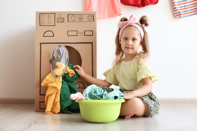 Adorable little child playing with cardboard washing machine and clothes indoors