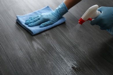 Photo of Woman in gloves cleaning black wooden table with rag and detergent indoors, closeup