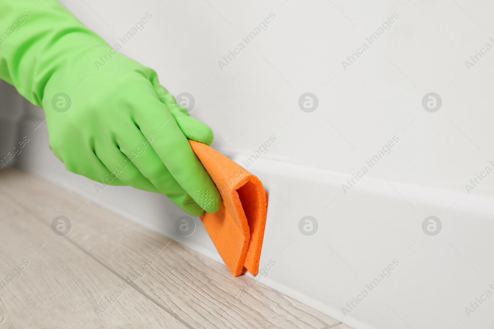 Photo of Woman in protective glove cleaning plinth with sponge cloth indoors, closeup. Space for text