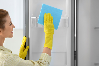 Photo of Woman in rubber gloves cleaning empty refrigerator with rag at home