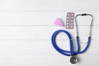 Flat lay composition with stethoscope, menstrual cup and pills on white wooden table, space for text. Gynecology concept