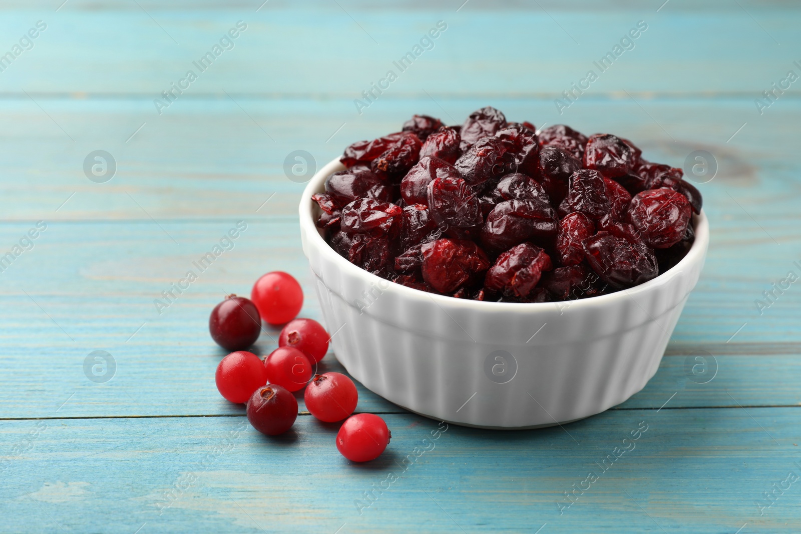 Photo of Tasty dried cranberries in bowl and fresh ones on light blue wooden table, closeup. Space for text