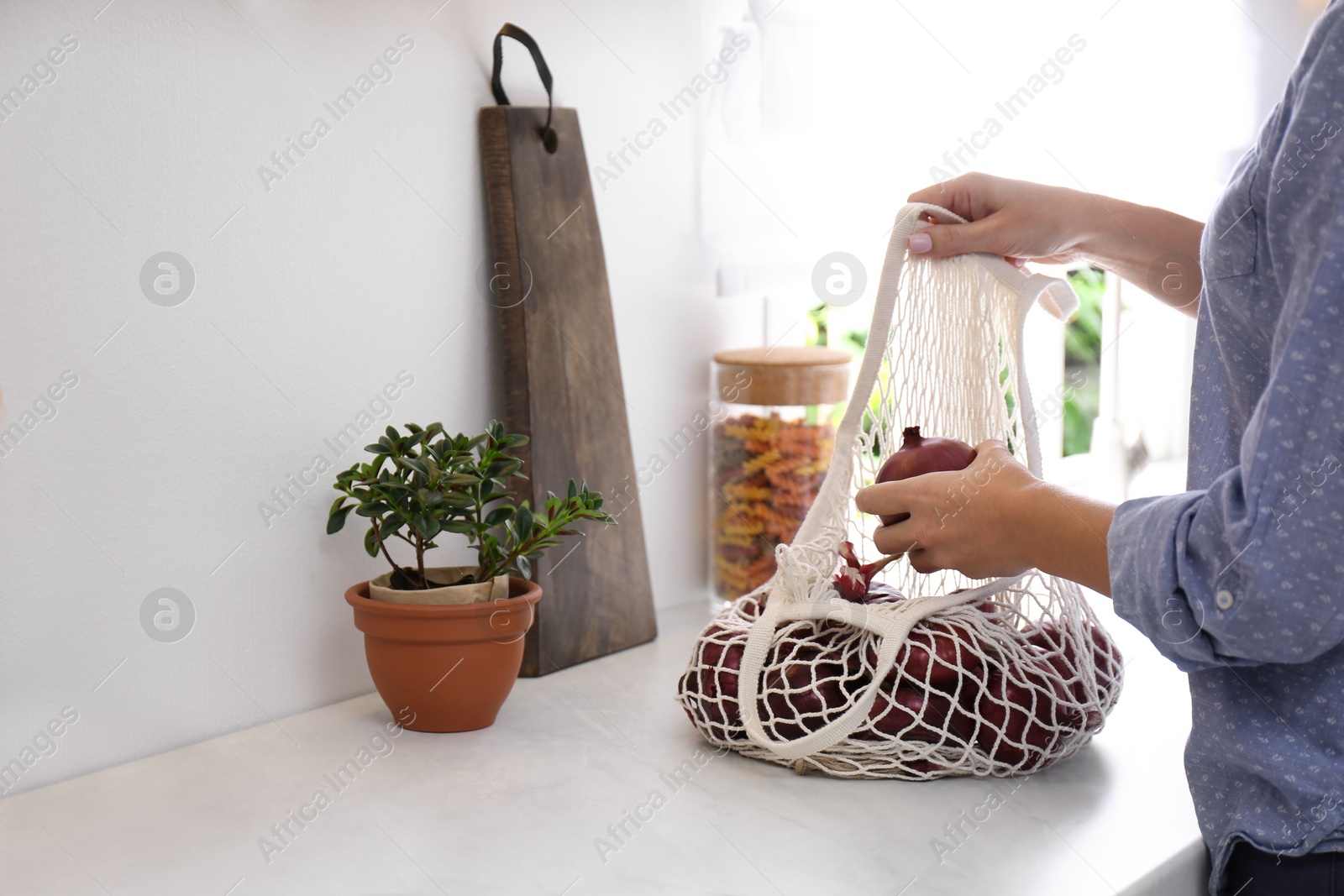 Photo of Woman taking red onion from mesh tote bag at countertop in kitchen, closeup