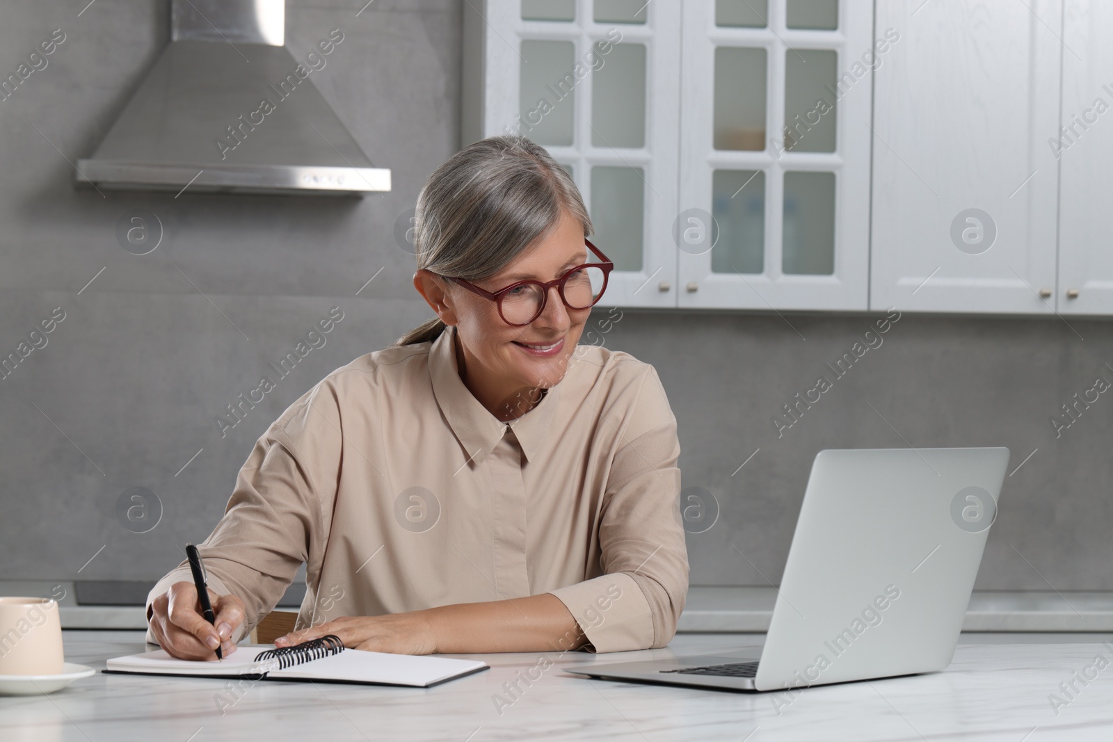 Photo of Beautiful senior woman taking notes while using laptop at white marble table in kitchen