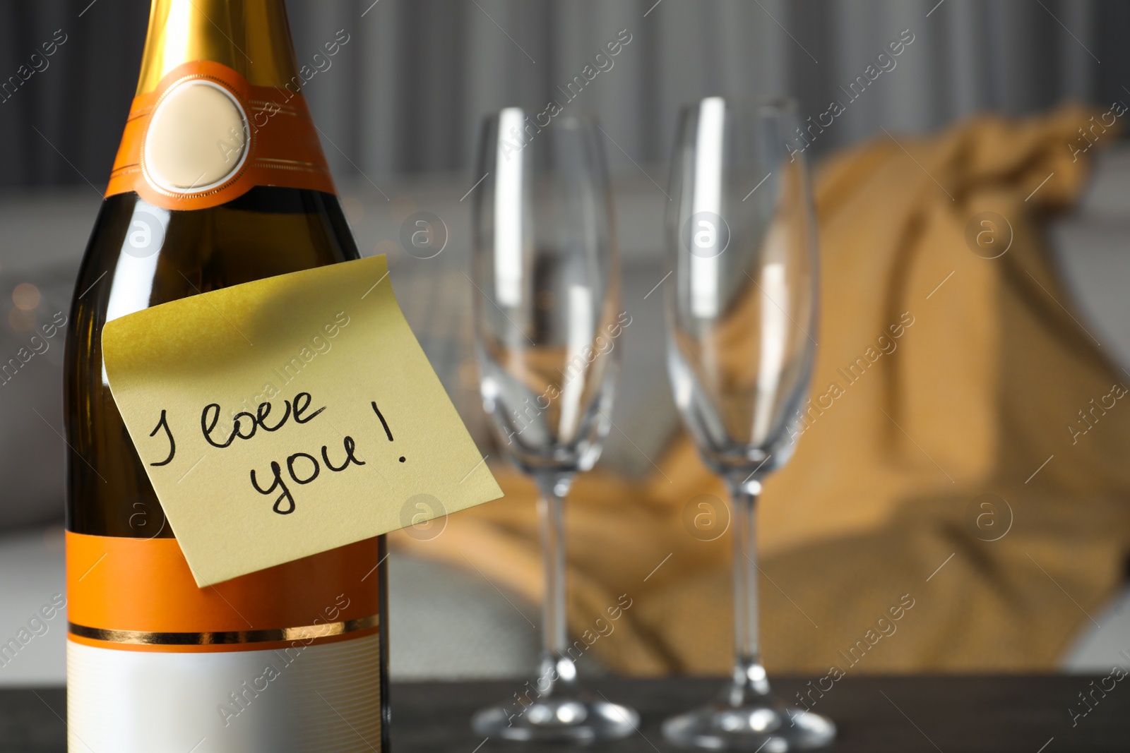 Photo of Note with phrase I Love You attached to bottle of champagne near glasses on table in room, closeup. Space for text