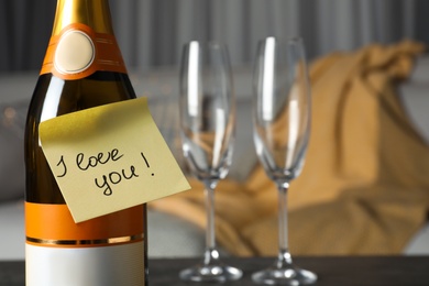 Photo of Note with phrase I Love You attached to bottle of champagne near glasses on table in room, closeup. Space for text