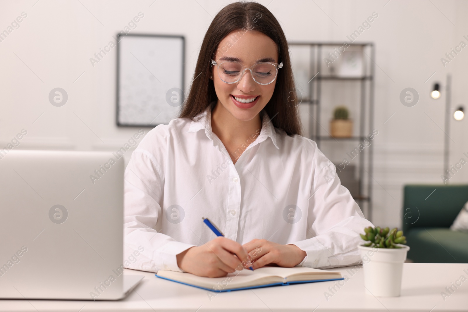 Photo of Young woman writing in notebook at white table indoors