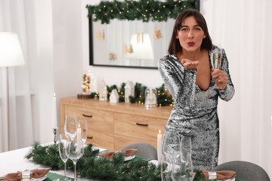 Photo of Beautiful woman holding glass of champagne and blowing kiss in room with Christmas decorations. Space for text
