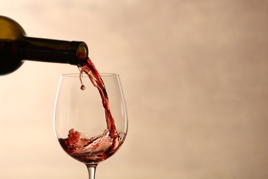 Pouring red wine into glass from bottle against blurred beige background, closeup. Space for text