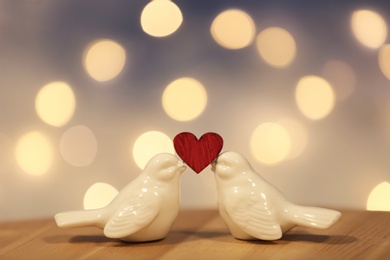 Photo of Composition with bird figures and small red heart on table against blurred lights