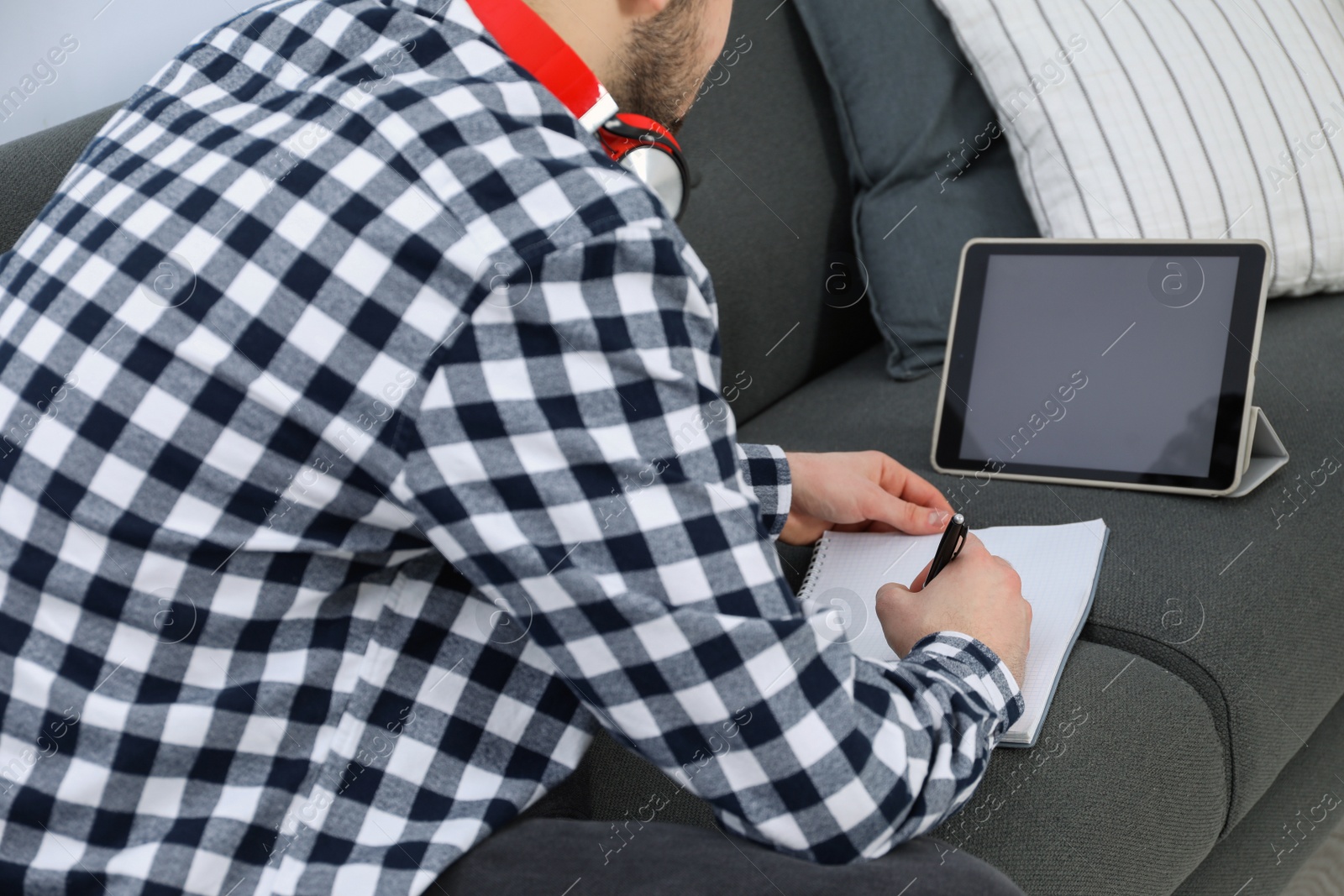 Photo of Young man with modern tablet and headphones studying on sofa, closeup. Distance learning