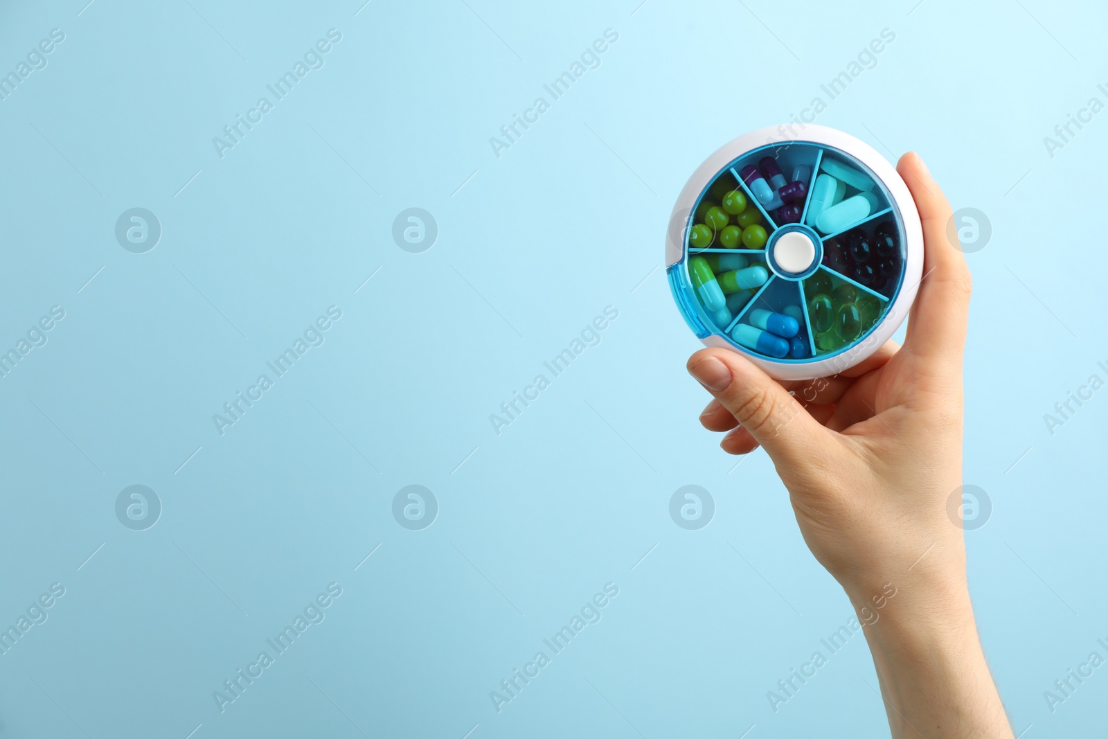 Photo of Woman holding plastic box with different pills on light blue background, closeup. Space for text
