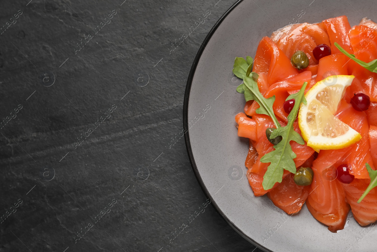 Photo of Salmon carpaccio with capers, cranberries, arugula and lemon on black table, top view. Space for text